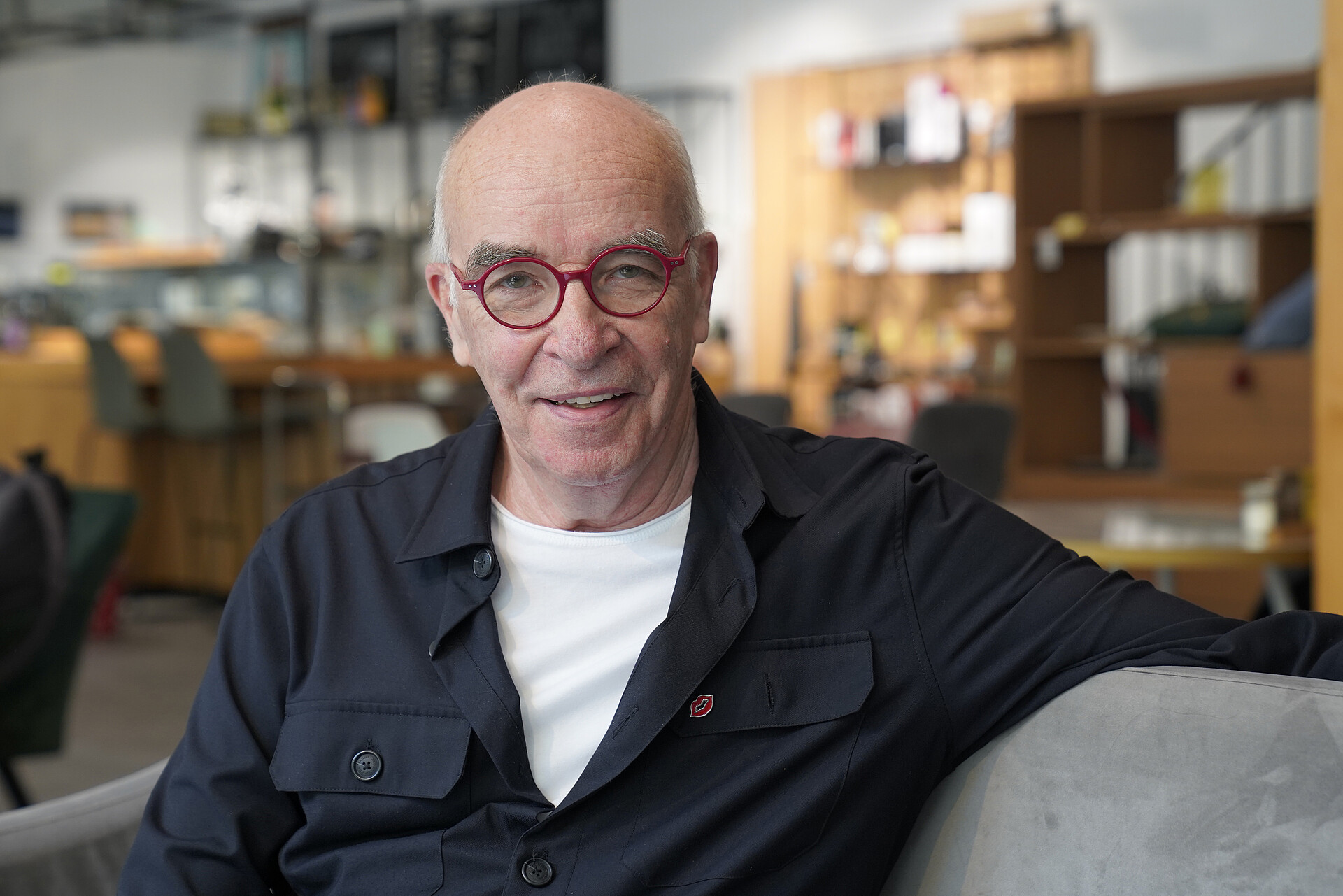 Helmut Geduldig in dunklem Hemd über weißem T-Shirt und roter Brolle auf einer Couch sitzend.