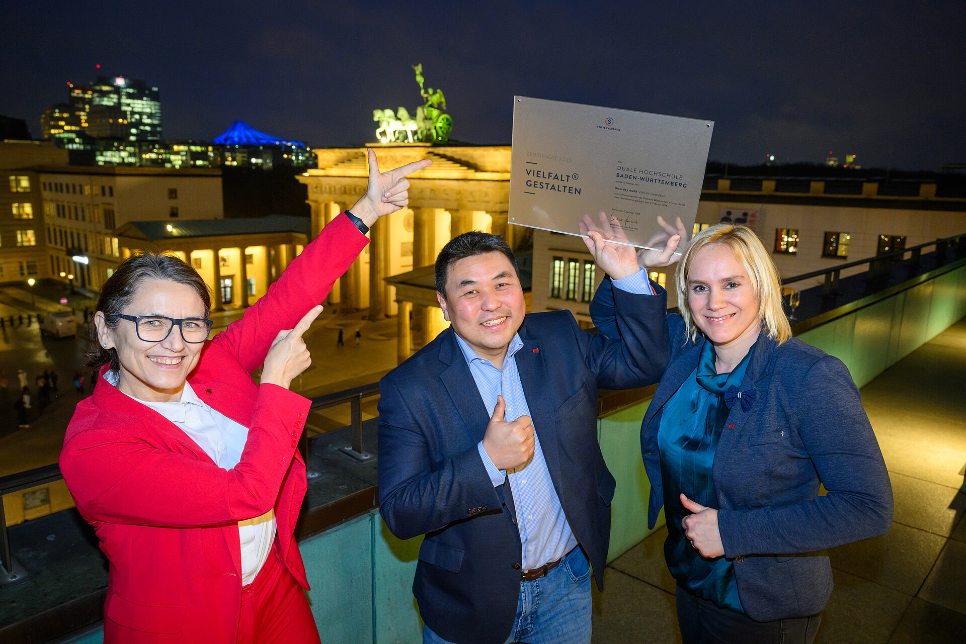 Von links nach rechts: Prof. Dr. Martina Klärle, Prof. Dr. Seon-Su Kim und Dr. Katharina Thomalla mit dem Zertifakt auf einem Balkon vor dem beleuchteten Brandenburger Tor.